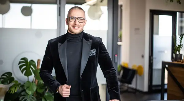 Mężczyzna w czarnym garniturze i okularach stoi w nowoczesnym biurze.