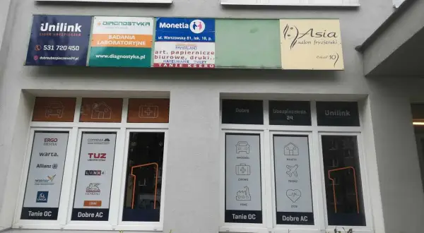 Szyldy firm ubezpieczeniowych i salonu fryzjerskiego na szarej ścianie budynku.