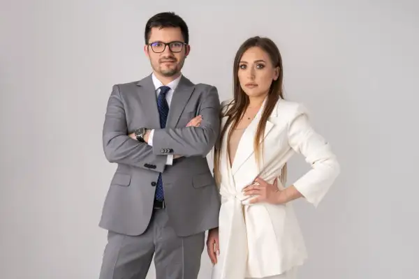 Para w garniturach, mężczyzna z rękami skrzyżowanymi na piersi, kobieta z ręką na biodrze.